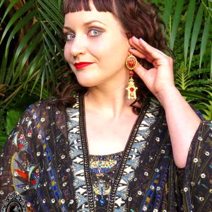 May include: A woman with dark brown hair is wearing a black and gold patterned kimono with a red and gold scarab beetle dangle earring. The earring has a red stone and gold accents.