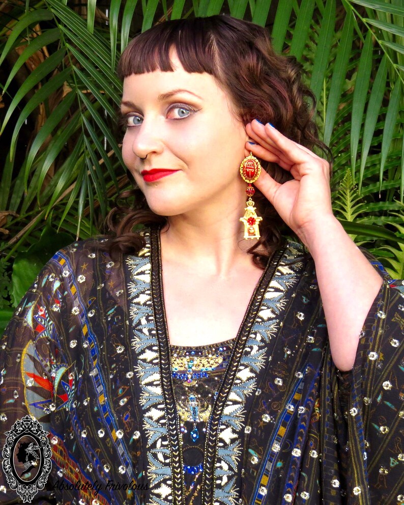 May include: A woman with dark brown hair is wearing a black and gold patterned kimono with a red and gold scarab beetle dangle earring. The earring has a red stone and gold accents.