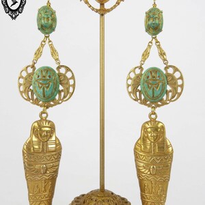 May include: A pair of gold-tone dangle earrings with green scarab beetle charms and a gold-tone sarcophagus charm. The earrings are displayed on a gold-tone stand.