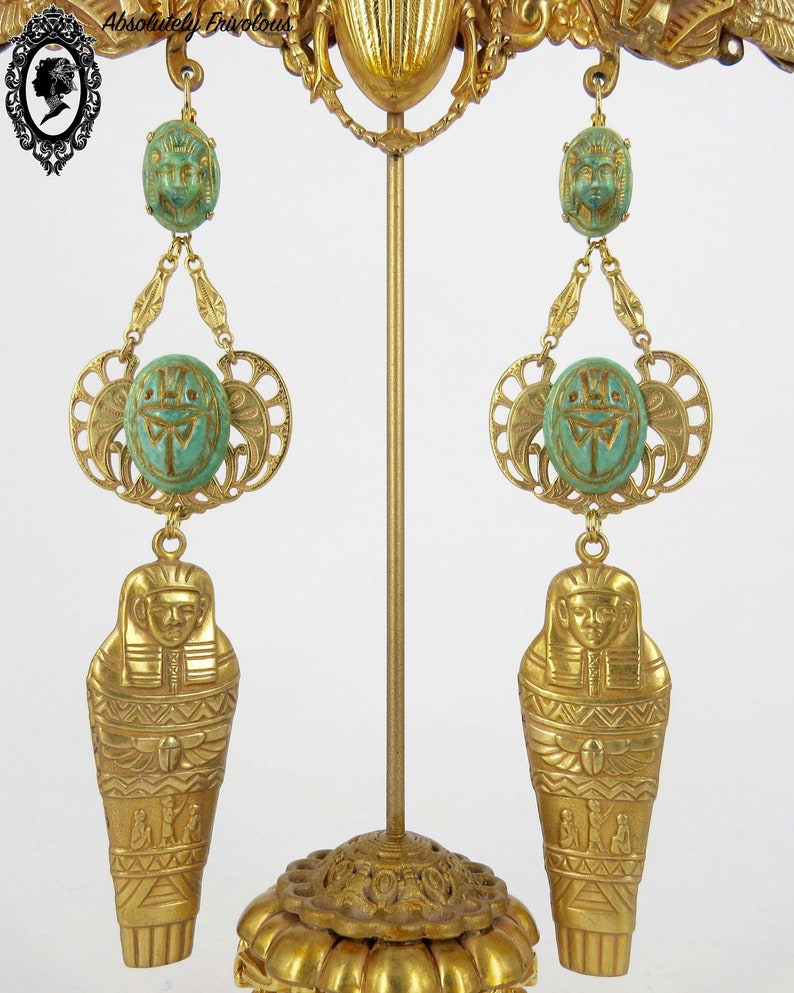 May include: A pair of gold-tone dangle earrings with green scarab beetle charms and a gold-tone sarcophagus charm. The earrings are displayed on a gold-tone stand.