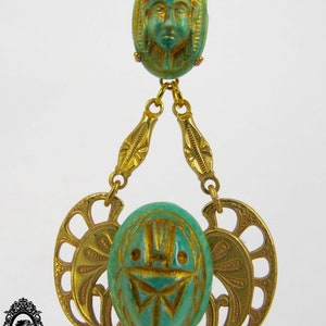May include: A gold chain necklace with two green and gold pendants. The top pendant features a carved face and the bottom pendant features a carved scarab beetle.