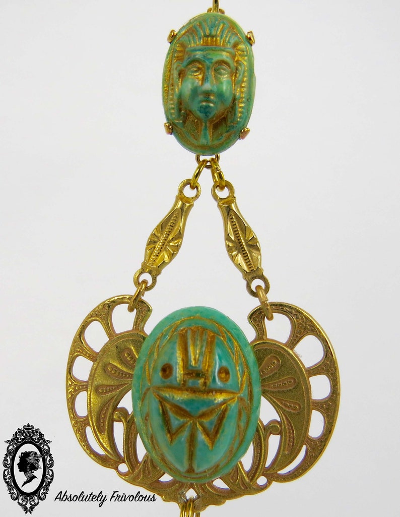May include: A gold chain necklace with two green and gold pendants. The top pendant features a carved face and the bottom pendant features a carved scarab beetle.