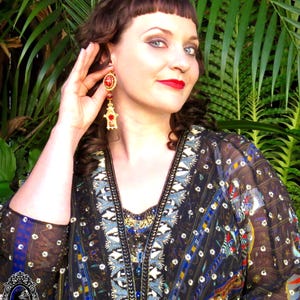 May include: A woman wearing a black and blue patterned blouse with gold and blue embroidery. She is wearing large red and gold dangle earrings.