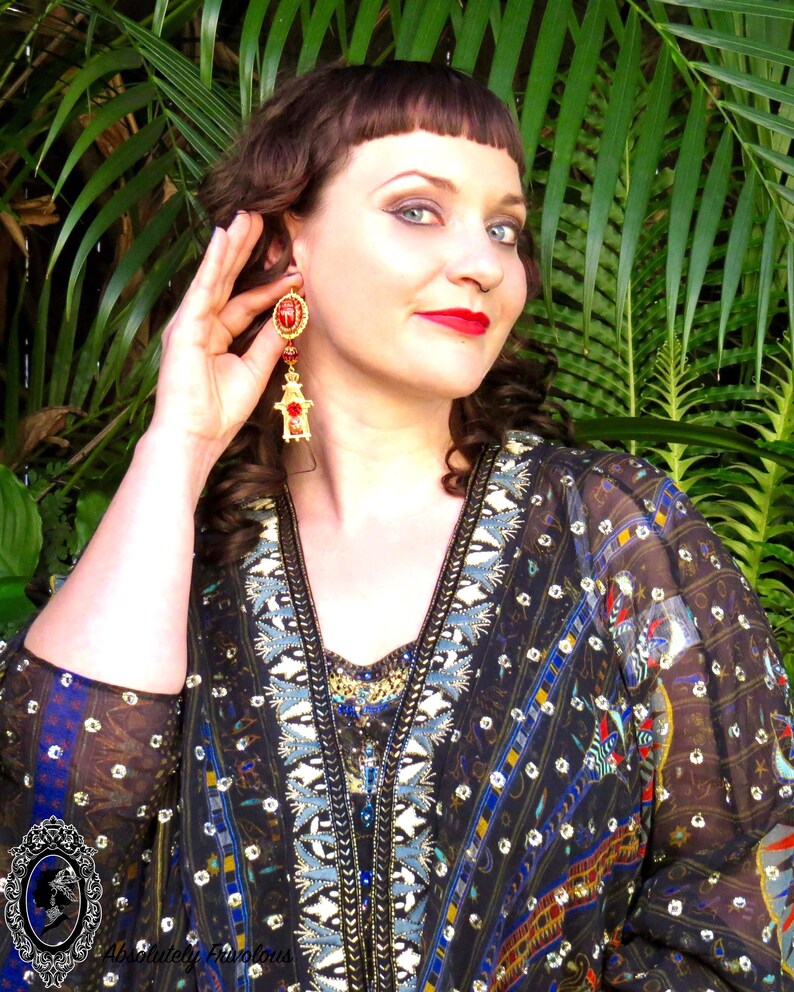 May include: A woman wearing a black and blue patterned blouse with gold and blue embroidery. She is wearing large red and gold dangle earrings.