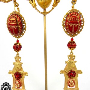 May include: Gold tone earrings with red beetle charms, red glass beads, and gold tone scarab figures. The earrings are displayed on a gold stand.