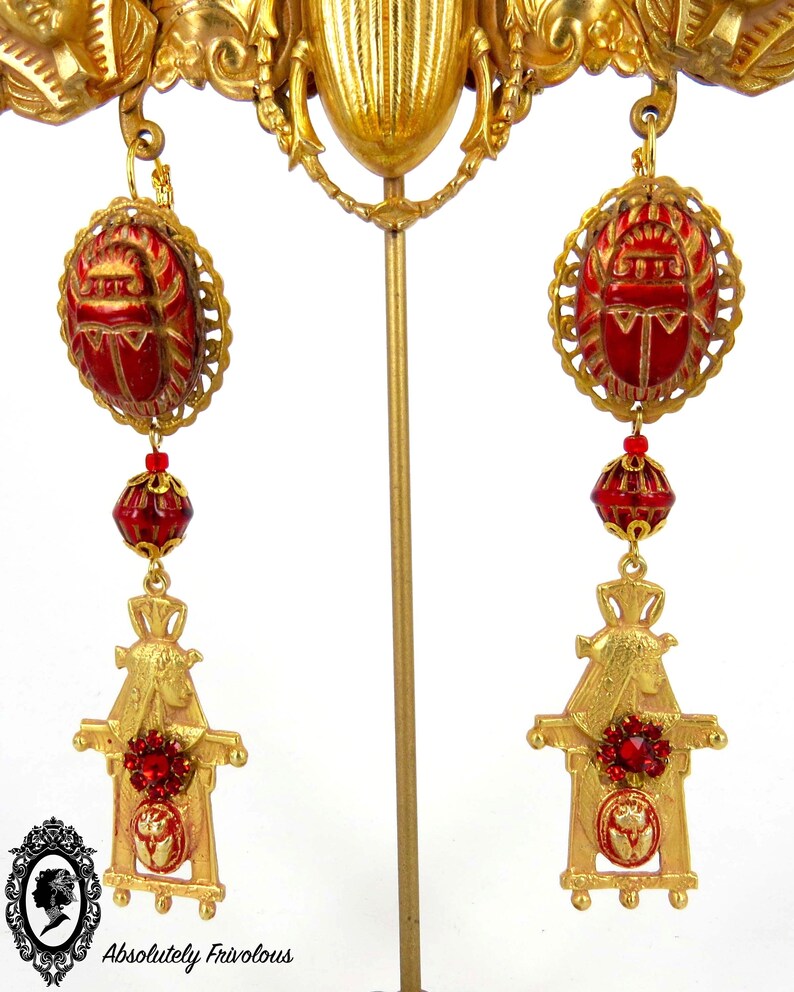 May include: Gold tone earrings with red beetle charms, red glass beads, and gold tone scarab figures. The earrings are displayed on a gold stand.