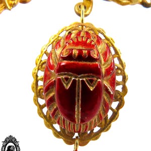 May include: A gold pendant with a red scarab beetle charm. The beetle is surrounded by a gold filigree design. The pendant is attached to a gold chain.