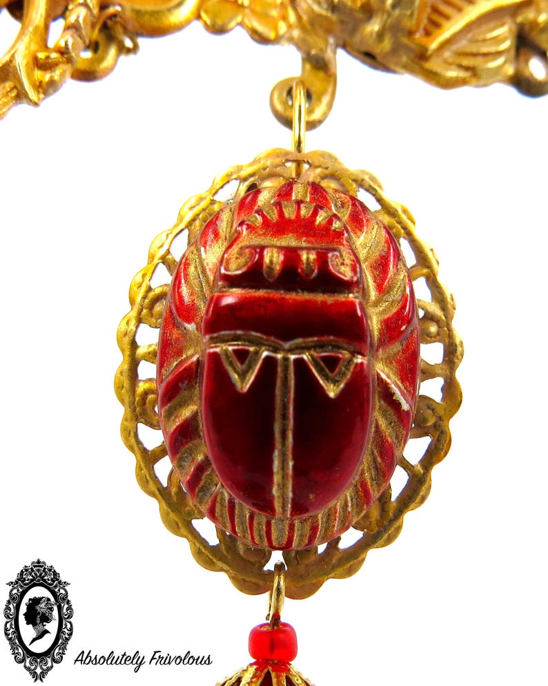 May include: A gold pendant with a red scarab beetle charm. The beetle is surrounded by a gold filigree design. The pendant is attached to a gold chain.