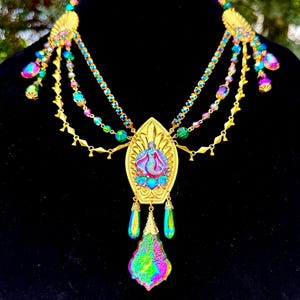 Op de afbeelding: Een goudkleurige statement ketting met een centrale hanger met een zeemeermin in blauw, roze en paars. De ketting heeft meerdere strengen kleurrijke kralen en iriserende kristallen accenten, met een grote, veelkleurige kristallen druppel.