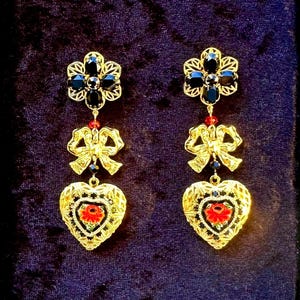 May include: Gold-toned drop earrings with a floral top, bow, and heart-shaped pendant. The earrings feature black gemstones, red and blue accents, and a red flower in the heart pendant. The earrings are ornate and detailed.