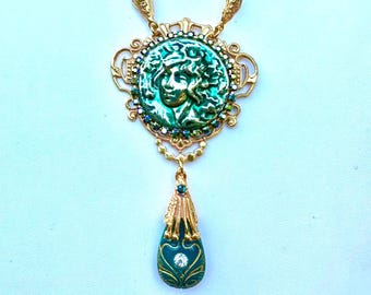 May include: A gold necklace with a green cameo pendant featuring a woman's face. The pendant is surrounded by gold filigree and green rhinestones. A smaller green teardrop pendant hangs from the bottom of the necklace.