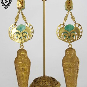 May include: Gold tone earrings with a green stone, a filigree design, and a dangling sarcophagus charm.  The earrings are displayed on a gold stand.