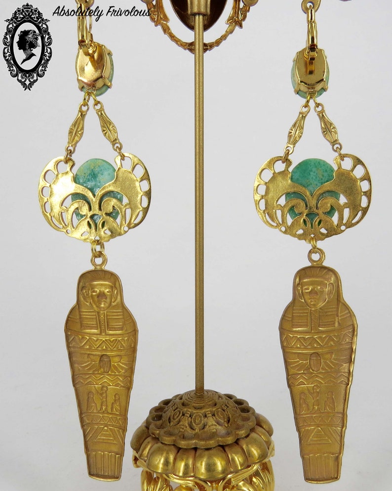 May include: Gold tone earrings with a green stone, a filigree design, and a dangling sarcophagus charm.  The earrings are displayed on a gold stand.