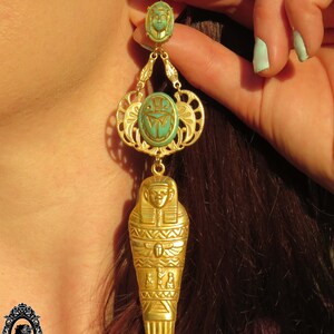 May include: Gold tone dangle earrings with a turquoise blue scarab beetle and a gold tone sarcophagus charm. The earrings are hanging from a woman's ear.