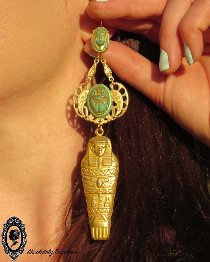 May include: Gold tone dangle earrings with a turquoise blue scarab beetle and a gold tone sarcophagus charm. The earrings are hanging from a woman's ear.