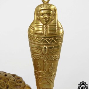 May include: Gold-tone pendant necklace featuring a detailed Egyptian sarcophagus design. The pendant is intricately carved with hieroglyphics and a depiction of a pharaoh.