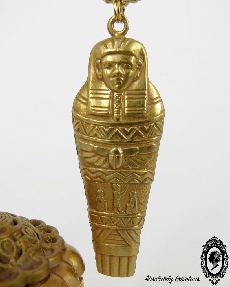 May include: Gold-tone pendant necklace featuring a detailed Egyptian sarcophagus design. The pendant is intricately carved with hieroglyphics and a depiction of a pharaoh.