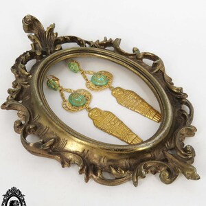 May include: A pair of gold-tone earrings with turquoise-colored stones and a sarcophagus design. The earrings are displayed in a gold-tone frame.
