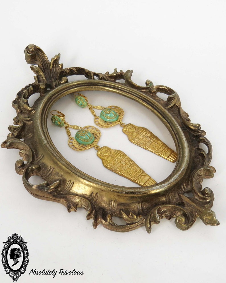 May include: A pair of gold-tone earrings with turquoise-colored stones and a sarcophagus design. The earrings are displayed in a gold-tone frame.