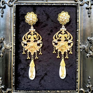 May include: A pair of ornate gold-colored earrings. Each earring features a floral-shaped top with clear crystals, a decorative face, and dangling pearl-like beads. The bottom of each earring has a white teardrop shape with gold accents. The earrings are displayed against a black background.