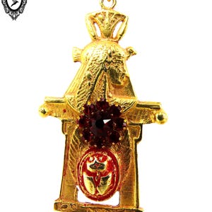 May include: Gold-tone pendant earring with a red jeweled center and a detailed design resembling a small building or structure.