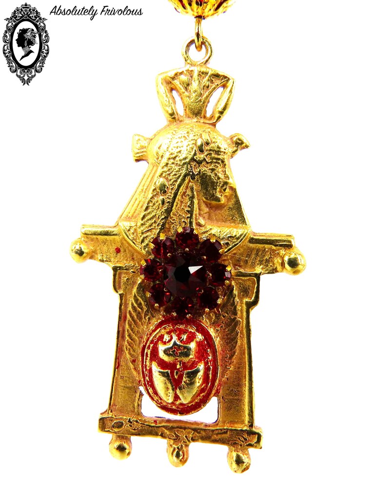 May include: Gold-tone pendant earring with a red jeweled center and a detailed design resembling a small building or structure.