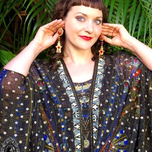 May include: A woman wearing a black and gold patterned kimono with red and gold dangle earrings. The kimono has a geometric pattern with blue, gold, and red accents. The earrings are shaped like a small building with a red roof.