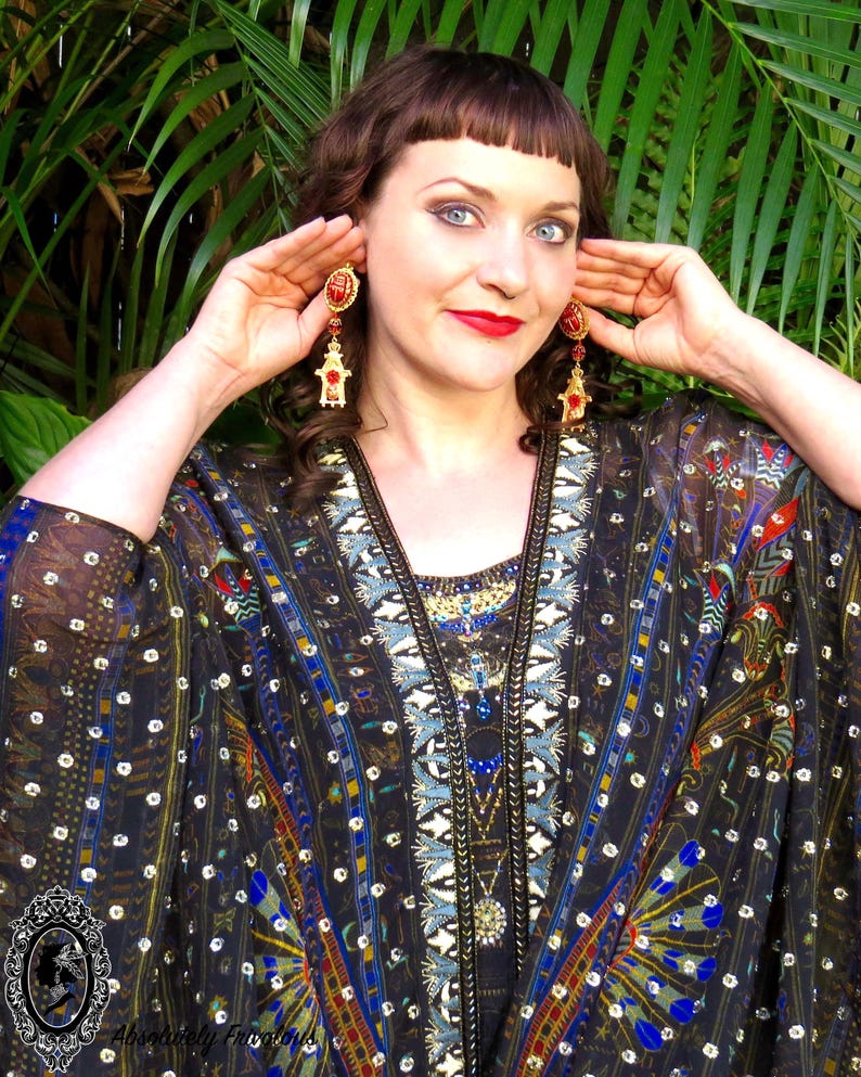 May include: A woman wearing a black and gold patterned kimono with red and gold dangle earrings. The kimono has a geometric pattern with blue, gold, and red accents. The earrings are shaped like a small building with a red roof.