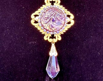 May include: A gold necklace with a purple cameo pendant featuring a woman's face surrounded by rhinestones. The pendant is attached to a teardrop-shaped crystal.