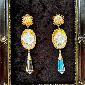 May include: A pair of ornate gold-toned earrings. Each earring features a floral top, an oval pendant with a figure, and a dangling, faceted crystal. One crystal is clear, the other is blue. The earrings are displayed against a black background.