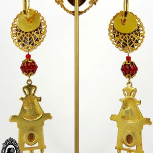 May include: Gold tone earrings with a red glass bead and a gold tone charm in the shape of a house. The earrings are hanging from a gold tone hoop.
