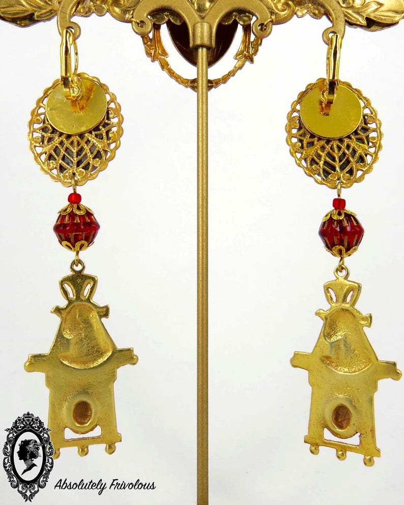 May include: Gold tone earrings with a red glass bead and a gold tone charm in the shape of a house. The earrings are hanging from a gold tone hoop.