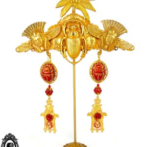 May include: Gold tone earrings with a red and gold scarab beetle design, hanging from a gold tone palm tree and winged figure design. The earrings are displayed on a gold tone stand.