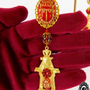 May include: Gold tone pendant necklace with a red scarab beetle charm, a red glass bead, and a gold tone temple charm with a red rhinestone.