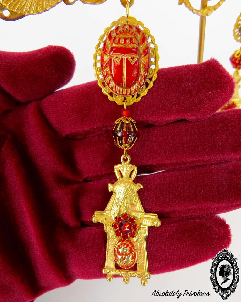 May include: Gold tone pendant necklace with a red scarab beetle charm, a red glass bead, and a gold tone temple charm with a red rhinestone.
