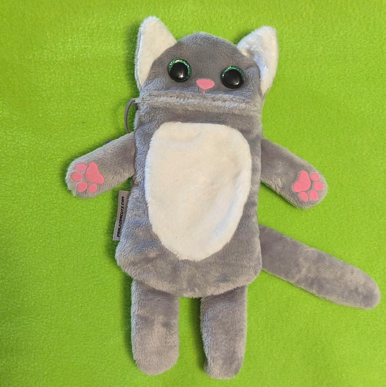 Puede incluir: Una bolsa de felpa gris con forma de gato con detalles blancos en el pecho, las orejas y la barriga. Tiene ojos grandes y brillantes, una nariz rosa y almohadillas en las patas. La bolsa tiene una cremallera y el texto "millasprouts.com".