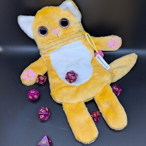 Puede incluir: Una bolsa de dados con forma de gato amarillo con detalles blancos y ojos grandes y brillantes. Huellas de patas rosas adornan las patas. Varios dados morados con n&uacute;meros blancos est&aacute;n esparcidos alrededor de la bolsa. Una etiqueta dice PROLACERUTZ.COM.