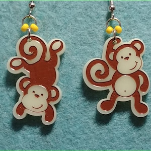 May include: A pair of brown monkey earrings with yellow beads. The monkeys are made of plastic and have a white background.
