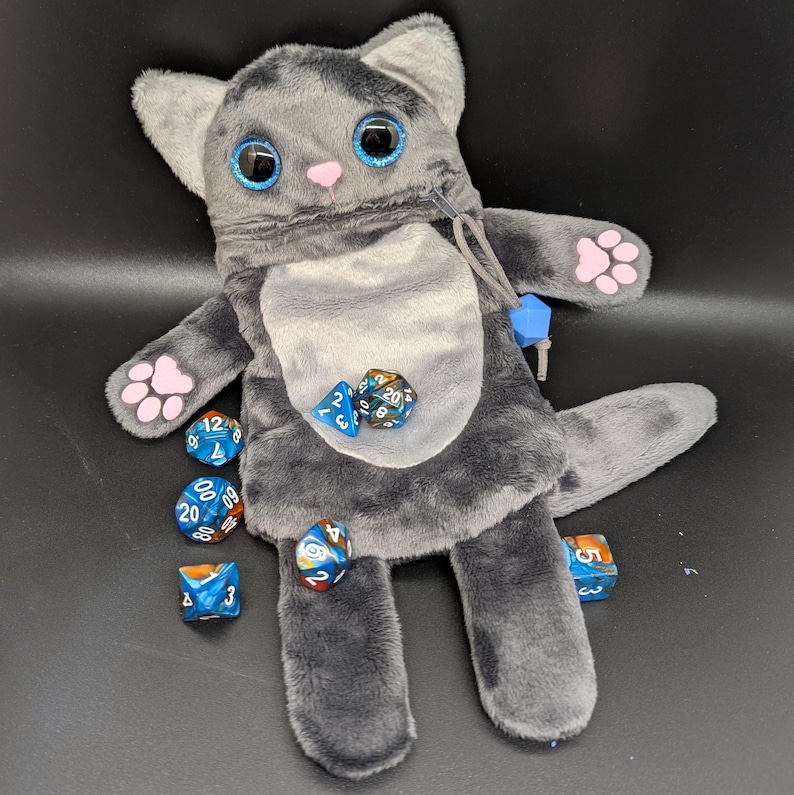 Puede incluir: Una bolsa de dados de felpa gris con forma de gato con ojos azules brillantes, nariz rosa y almohadillas para las patas. Varios dados multicolores est&aacute;n esparcidos alrededor de la bolsa. La bolsa tiene un bolsillo con cremallera y un cierre de cord&oacute;n.