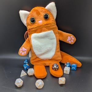 Puede incluir: Bolsa de dados de felpa naranja con forma de gato con detalles blancos, almohadillas rosas y ojos grandes y brillantes. Varios dados poli&eacute;dricos de diferentes colores y dise&ntilde;os est&aacute;n esparcidos alrededor de la bolsa, para juegos de mesa.