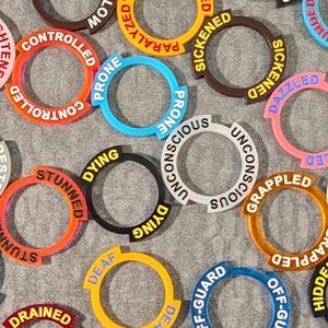 May include: A collection of colorful, circular tokens with various status effect labels like "Controlled," "Prone," and "Dying." The tokens are in shades of red, blue, yellow, and black, with white lettering. These tokens are likely used in tabletop gaming.