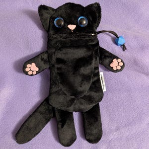 Puede incluir: Un objeto negro con forma de gato de peluche con grandes ojos azules y nariz y almohadillas rosas. El gato tiene una cremallera y un cord&oacute;n negro con una cuenta azul. El objeto est&aacute; sobre una superficie p&uacute;rpura.