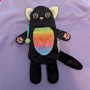 Puede incluir: Una bolsa con cremallera con forma de gato negro de peluche con una barriga de color arco&iacute;ris. El gato tiene ojos grandes y brillantes, nariz rosa y almohadillas en las patas. Tiene puntas de orejas blancas y un tirador de cremallera azul con un adorno geom&eacute;trico verde.