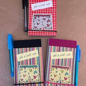 May include: Three small notebooks with underwear designs and the text "just a brief note." One notebook has a red grid pattern, while the other two have colorful striped patterns. Each notebook is secured with a black elastic band and accompanied by a pen.