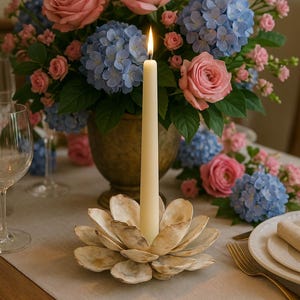 Könnte beinhalten: Eine brennende elfenbeinfarbene Kerze in einem muschelförmigen Halter auf einem Tisch, mit einem Blumenarrangement aus rosa Rosen und blauen Hortensien in einer goldenen Vase. Der Text "TONKIN HANDICRAFT" ist sichtbar.