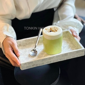 Puede incluir: Una bandeja rectangular blanca con un patrón decorativo de ladrillos sostiene un vaso de bebida verde cubierto con espuma y granola, y una cuchara pequeña. La bandeja tiene el texto "TONKIN HAND".