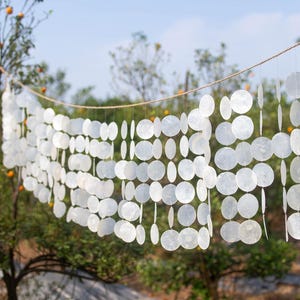 Op de afbeelding: Een decoratieve reeks witte, cirkelvormige schelpstukken hangt aan een touw, waardoor een gordijnachtig effect ontstaat. De tekst "TONKIN HANDICRAFT" is zichtbaar in het midden bovenaan. De achtergrond toont een blauwe lucht en een boom met sinaasappels.