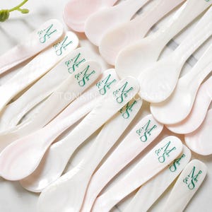 Könnte beinhalten: Eine Sammlung von weißen Perlmuttlöffeln mit grüner Beschriftung "Mr. & Mrs.". Die Löffel sind fächerförmig auf einer weißen Oberfläche angeordnet.