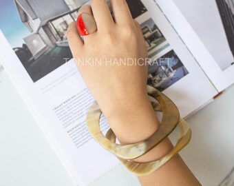 May include: A pair of brown horn bracelets with a red and brown ring on a hand. The hand is holding a book with the text "Tonkin Handicraft" on the page.