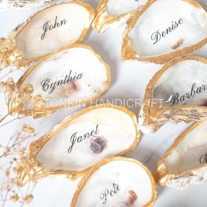 Puede incluir: Una colección de conchas de ostra con bordes pintados en oro, cada una con un nombre único escrito en letra negra. El interior de las conchas es blanco y liso, mostrando nombres como Tracey, Erin, John, Denise, Cynthia, Barbara, Janet y Pete.
